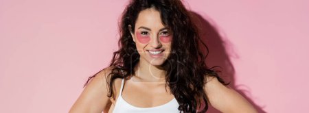 Smiling and curly woman with hydrogel eye patches looking at camera on pink background, banner 