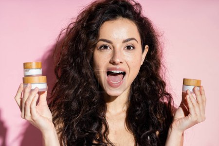 Excited curly woman holding jars with creams on pink background 