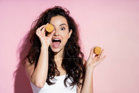 Shocked curly woman holding cosmetic creams on pink background 