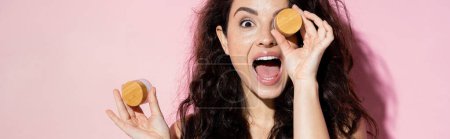 Photo for Shocked young woman holding jars with cosmetic creams on pink background, banner - Royalty Free Image