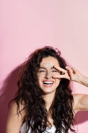 Cheerful curly woman showing victory sign on pink background 