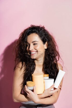 Cheerful curly woman holding cosmetic products on pink background 