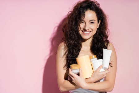 Positive curly woman holding beauty products on pink background 