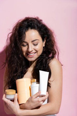 Positive brunette woman looking at cosmetic products on pink background 