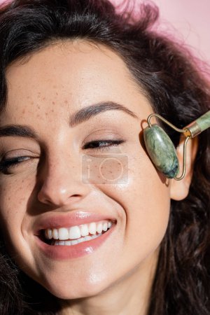 Téléchargez les photos : Close up view of smiling freckled woman using jade roller isolated on pink - en image libre de droit