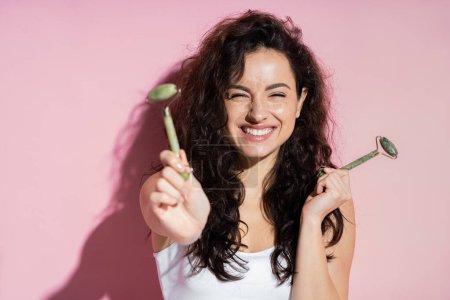 Positive freckled woman holding jade rollers on pink background 