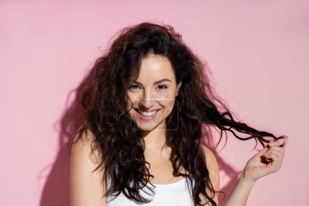 Téléchargez les photos : Cheerful curly woman in top touching hair on pink background - en image libre de droit