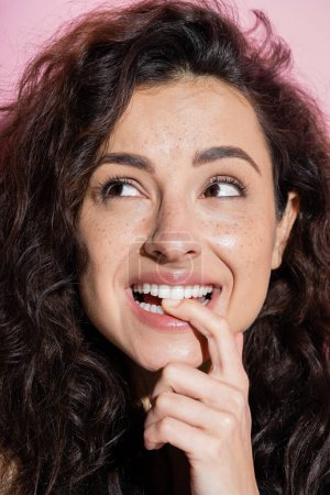 Pretty brunette woman with freckles holding finger near teeth isolated on pink 