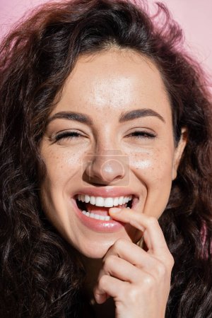 Positive curly woman looking at camera and holding finger near teeth isolated on pink 