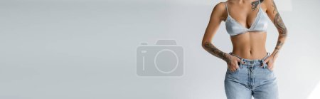 Téléchargez les photos : Cropped view of slender woman in satin bra standing with hands in pockets of jeans on grey background, banner - en image libre de droit