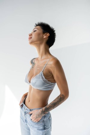 seductive tattooed woman in silk bra posing with hands in pockets of jeans on grey background