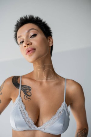 young woman with sexy tattooed body wearing blue satin bra and looking at camera on grey background