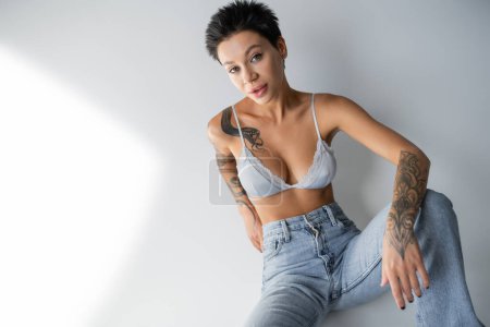 brunette woman with sexy tattooed body sitting in jeans and bra on grey background