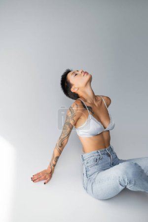 seductive tattooed woman in silk bra and jeans sitting with closed eyes on grey background