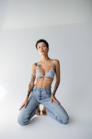 sensual brunette woman in satin bralette and jeans posing on knees on grey background