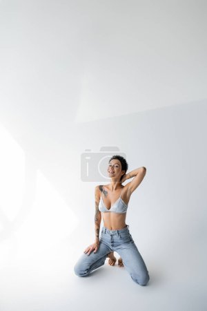 happy tattooed woman in jeans and bra touching neck while posing on knees on grey background
