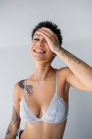 cheerful tattooed woman in blue silk bra obscuring face with hand isolated on grey
