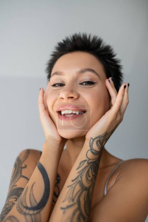 portrait of cheerful tattooed woman with short hair and piercing holding hands near face on grey background