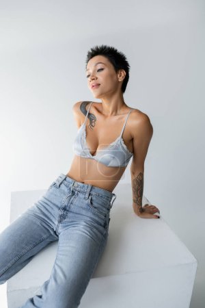 young tattooed woman in blue jeans and satin bra posing on cube on grey background Poster 638592362