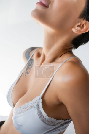 partial view of sexy tattooed woman in blue silk bralette on grey background