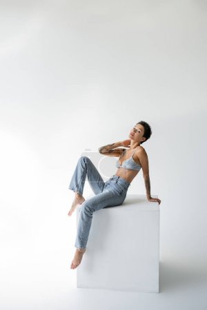 young barefoot woman in blue jeans and satin bra posing on cube and looking at camera on grey background
