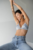 joyful tattooed woman in jeans and silk bra sitting on cube with raised hands and closed eyes on grey background tote bag #638592494