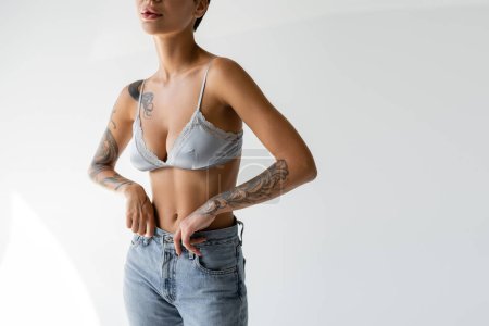 Téléchargez les photos : Cropped view of tattooed and slim woman posing in silk bra and blue jeans on grey background - en image libre de droit