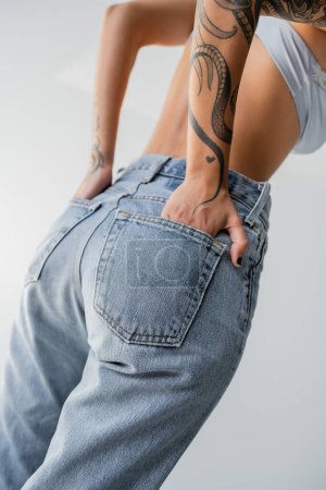 Téléchargez les photos : Cropped view of tattooed woman in bra posing with hands in back pockets of blue jeans isolated on grey - en image libre de droit