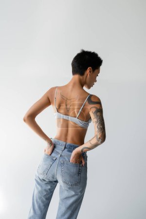 back view of tattooed woman in bra holding hands in back pockets of jeans on grey background