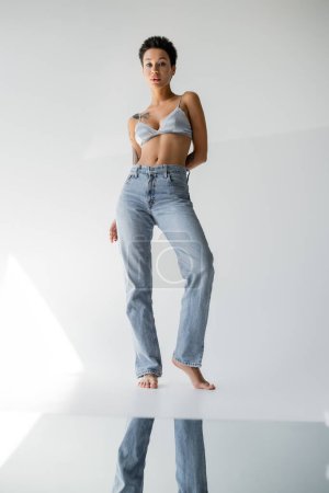 full length of barefoot brunette woman in jeans and bralette standing near mirror on floor on grey background