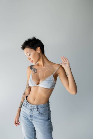 seductive brunette woman touching strap of blue bralette while posing in blue jeans isolated on grey
