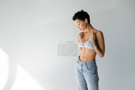 sexy woman with short brunette hair wearing blue jeans and touching straps of satin bra on grey background