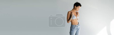 slender tattooed woman in blue jeans touching straps of silk bralette on grey background, banner