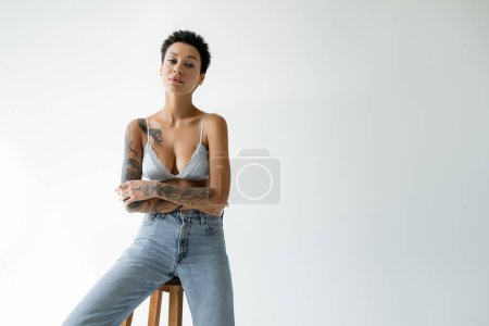Photo for Young tattooed woman in blue bralette and jeans sitting on stool on grey background - Royalty Free Image