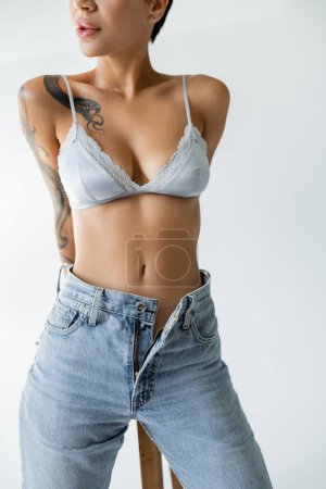 partial view of seductive woman in silk bra and unzipped jeans standing with hands behind back on grey background