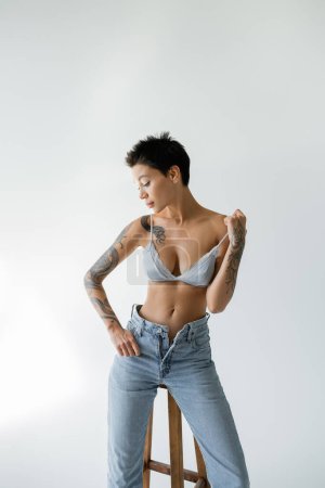 tattooed woman in unzipped jeans and silk bra posing near high stool on grey background