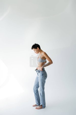 Téléchargez les photos : Longueur totale de pieds nus et femme mince en soutien-gorge en soie et jeans dézippés debout sur fond gris - en image libre de droit