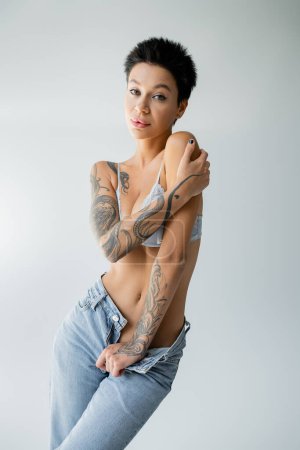 sensual tattooed woman in bra and unzipped jeans looking at camera isolated on grey