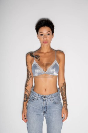 front view of tattooed woman with short hair wearing blue silk bra and jeans on grey background