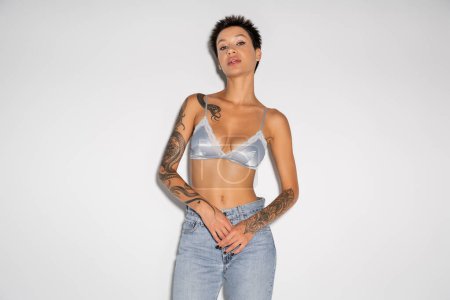 front view of brunette tattooed woman in silk bra and blue jeans looking at camera on grey background