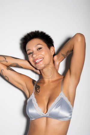 happy tattooed woman with short brunette hair posing in blue satin bra on grey background