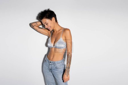Foto de Smiling and sexy woman in silk bralette and jeans standing with hand behind head on grey background - Imagen libre de derechos