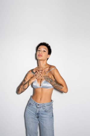 sexy tattooed woman in silk bra and jeans holding hands on chest while looking at camera on grey background