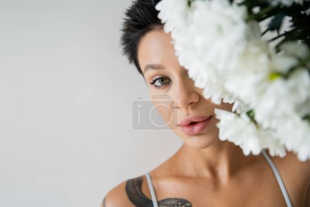 Foto de Retrato de mujer tatuada con maquillaje que oscurece la cara con flores blancas y mirando a la cámara aislada en gris - Imagen libre de derechos