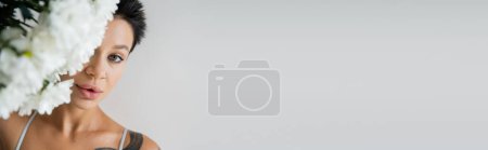 Téléchargez les photos : Portrait de femme brune maquillée regardant caméra près de fleurs blanches sur fond flou isolé sur gris, bannière - en image libre de droit