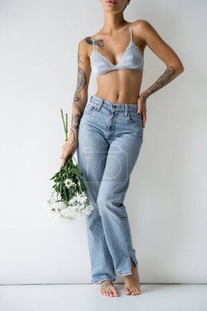 Photo for Partial view of tattooed barefoot woman in silk bra and jeans standing with hand on hip and white flowers on grey background - Royalty Free Image