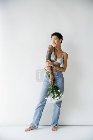 full length of barefoot woman in silk bra and jeans holding bouquet and looking away on grey background