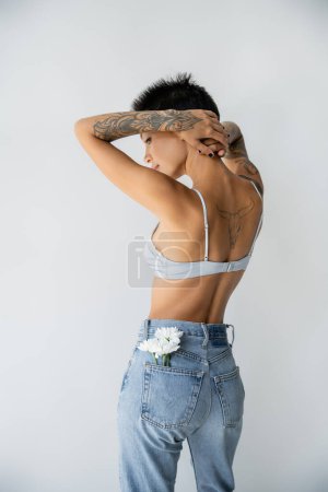 tattooed woman in bra and jeans with white flowers in back pocket standing with hands behind head isolated on grey