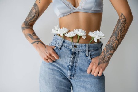 cropped view of tattooed woman in silk bralette and jeans with white flowers standing with thumbs in pockets isolated on grey