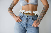 cropped view of tattooed woman in silk bralette and jeans with white flowers standing with thumbs in pockets isolated on grey Stickers #638593508